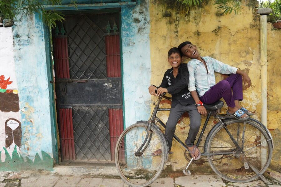 Indian children smiling on bicycles smiling at camera in front of yellow wall