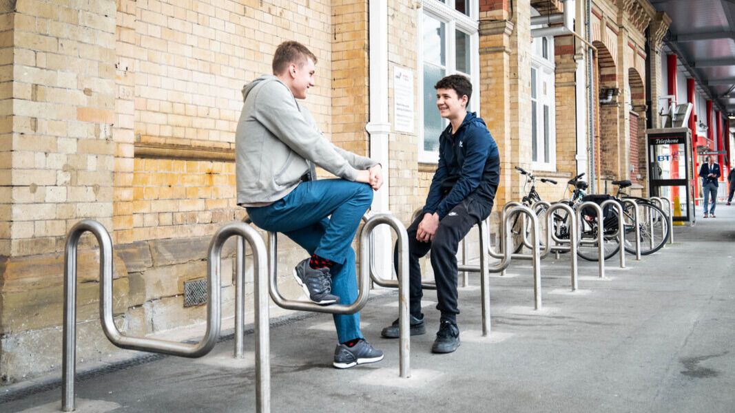 Two teenage boys sat talking to each other at a train station.