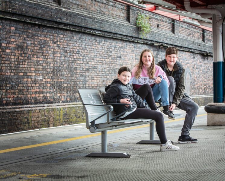 Three young teenagers sitting on a bench at a railway station in the UK.