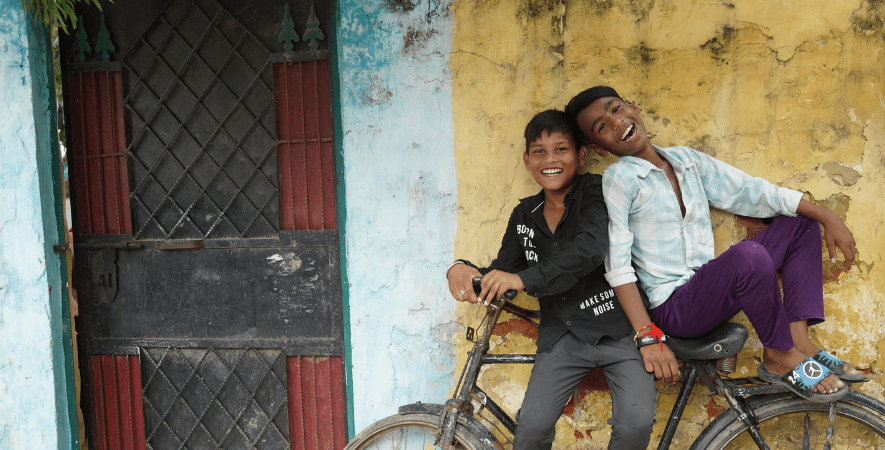 Two boys in India, one is leaning against a bike and the other is sitting on it. They are both smiling.