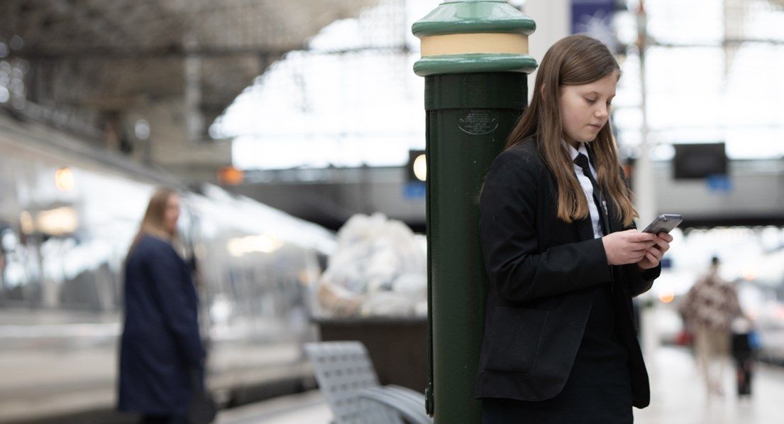 Young girl in school uniform on phone at a train station, looking vulnerable.