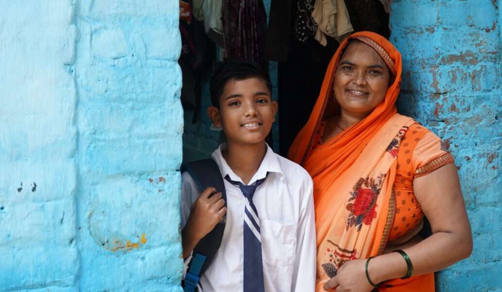 Indian boy in school uniform and mother in orange sari stand in doorway smiling at camera