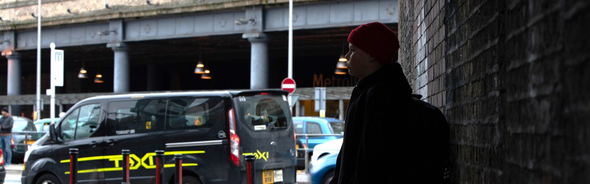 Teenage boy with a red hat carrying a backpack and leaning against a wall looking into the distance, a taxi is passing by on a nearby road.