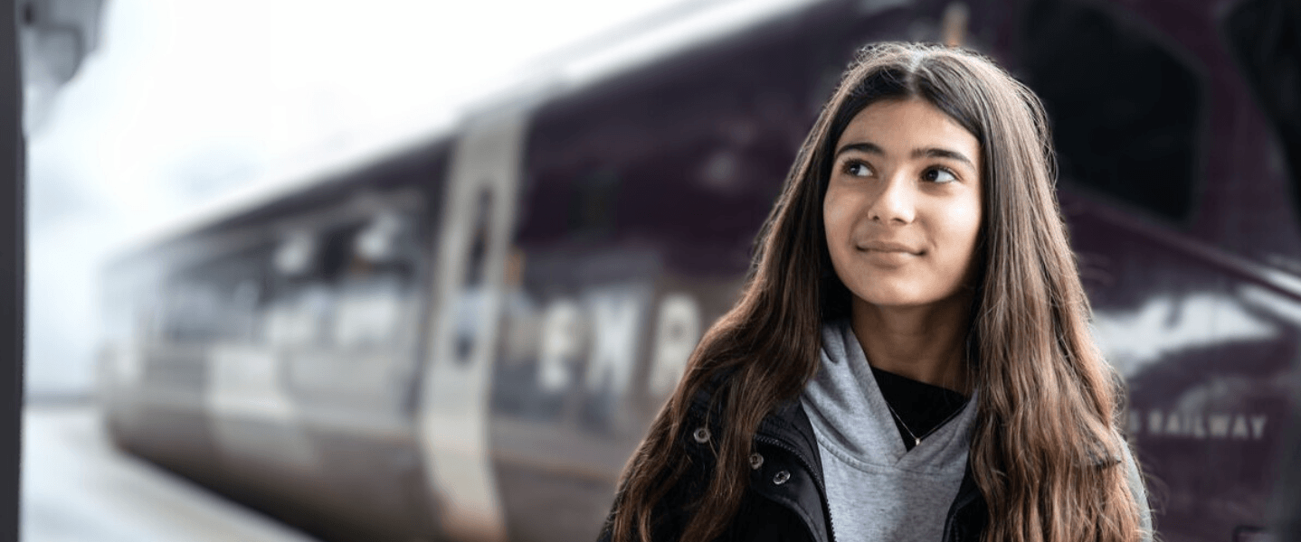 Young teenage girl stood in front of the side of a train smiling and looking into the distance on a train station platform in the UK